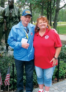 An older man wearing a blue Vietnam veteran hat and jacket poses with a red-headed woman in a red Honor Flight Network shirt in front of a green and black memorial statue of three Vietnam soldiers.