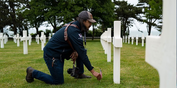 Man in baseball cap kneels, placing an American flag at a gravesite with a white cross headstone