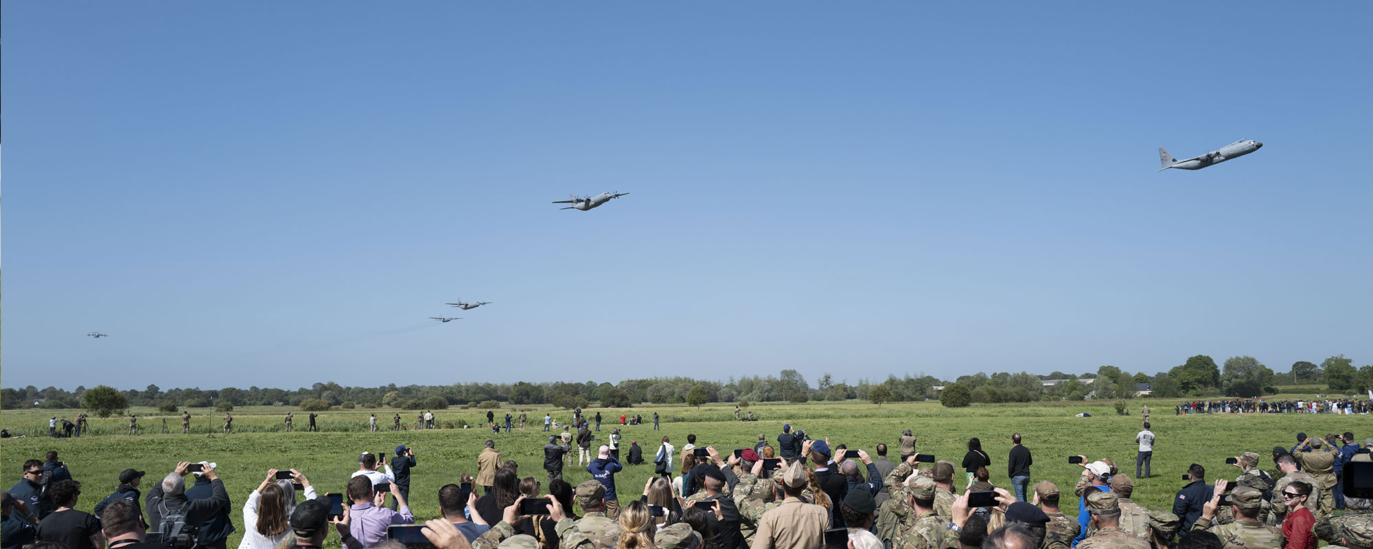 A crowd of people on a green grass airfield photograph five planes flying overhead