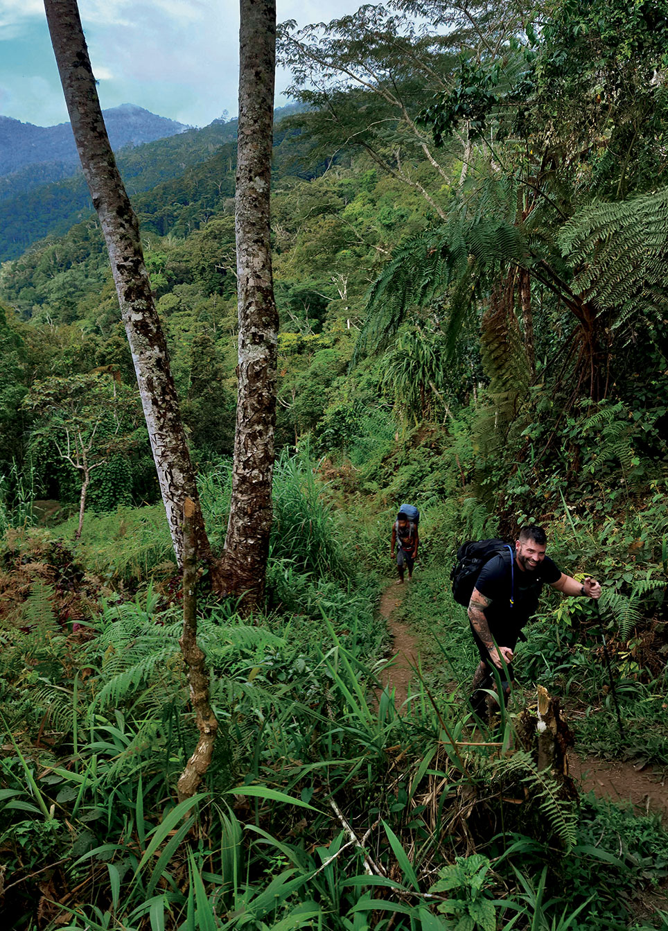 Man in black t-shirt using hiking sticks to climb a steep trail in a tropical jungle.