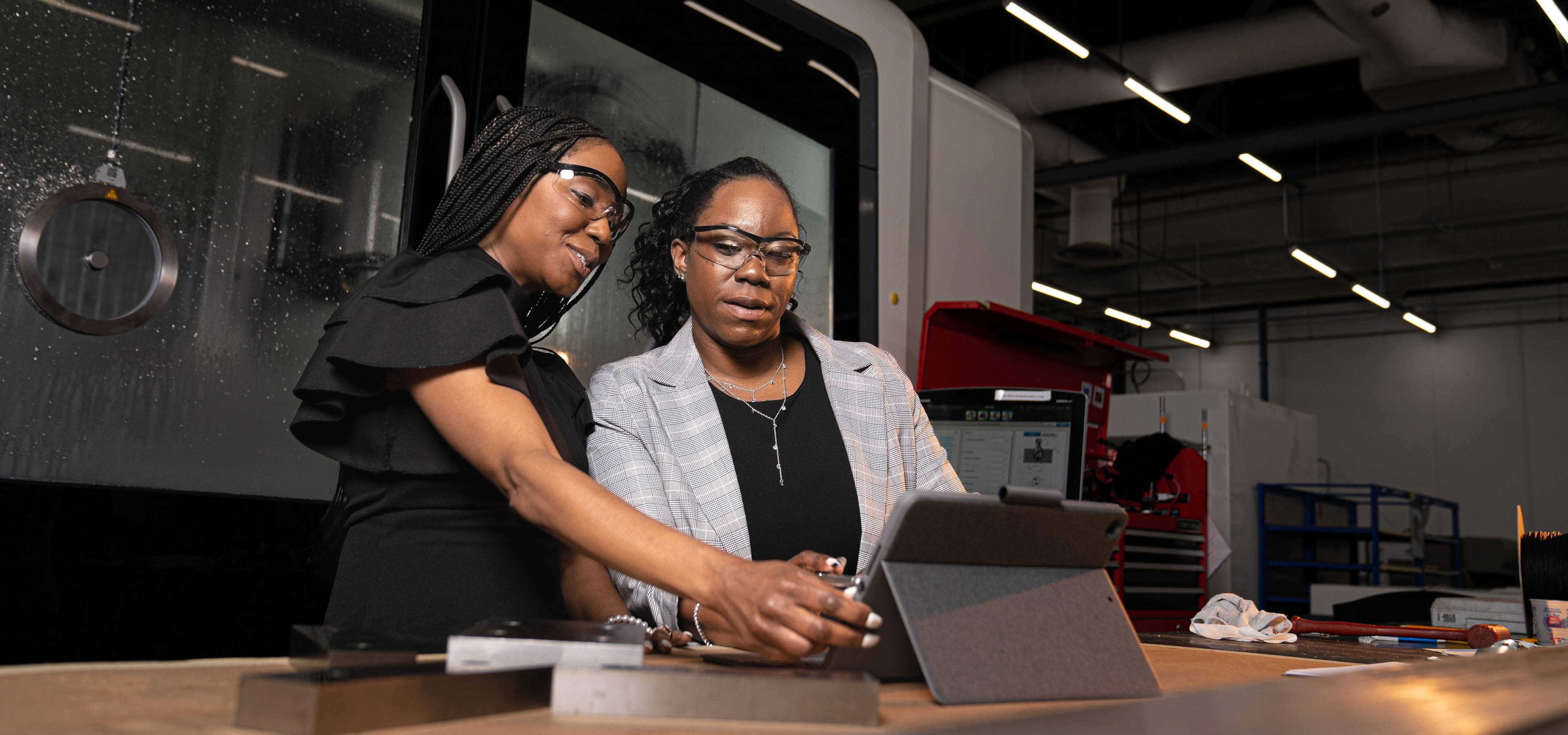Two women in a manufacturing setting wear safety goggles while reviewing an iPad.