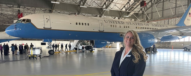 A blonde woman in a navy suit stands in front of a white plane in a hangar.