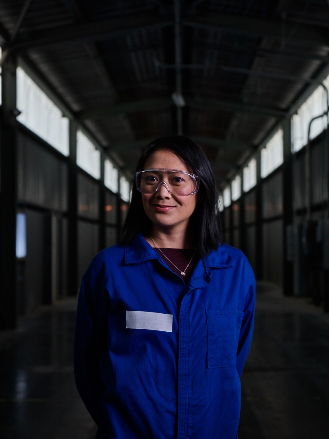 Asian female in blue lab coat and safety goggles stands in hallway of manufacturing plant