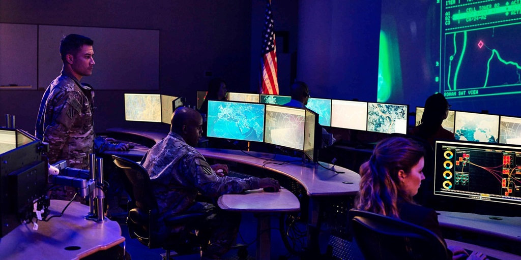 5 people in dark lit control room looking at computer screens dressed in military camouflaged suits and business attire