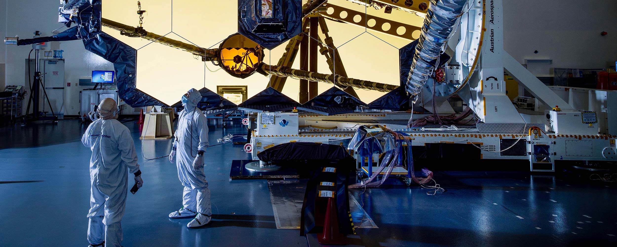 Two people in clean suits look up at the James Webb Space Telescope