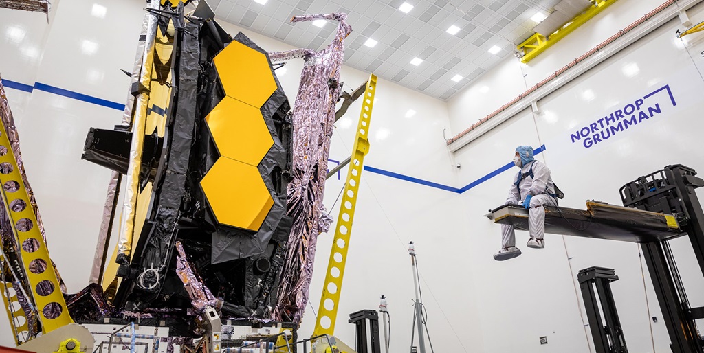 An engineer in a clean suit sits on a crane and looks at the James Webb Space Telescope sunshield