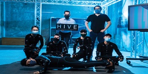 Seven engineers pose for a picture in a blue lit room. Five of them are in black jump suits.