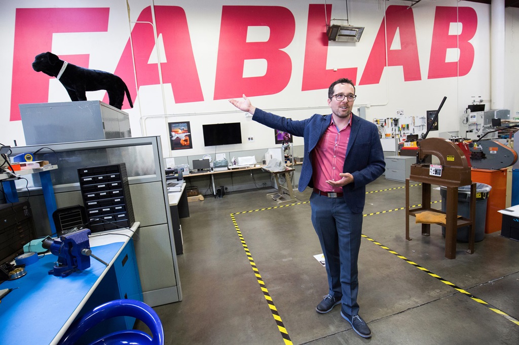 A man in a blue suit stands in a large workroom with the word "FabLab" painted on the wall behind him