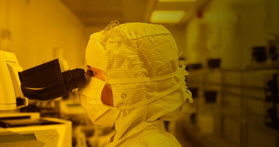 woman in clean room wearing full bunny suit looking through microscope