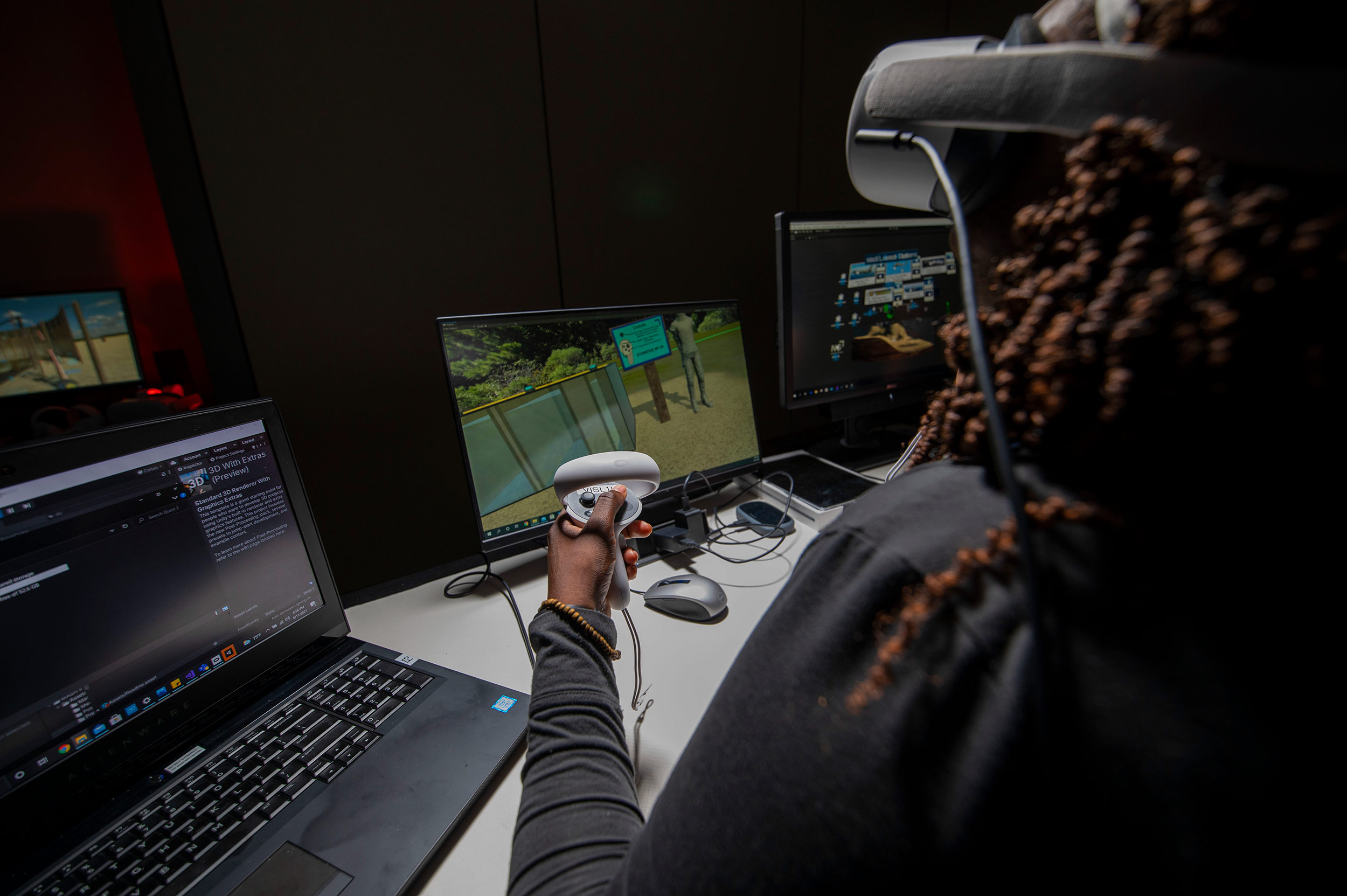 A woman sits at a computer wearing a virtual reality headset and holding a game controller