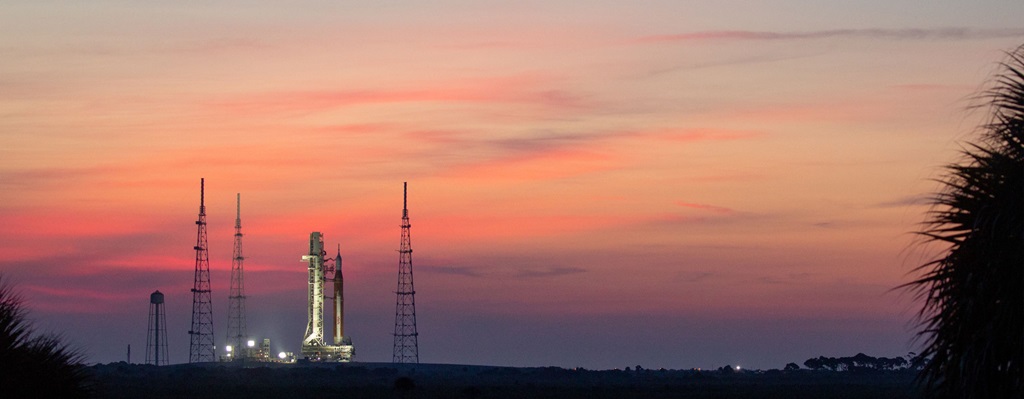View of Artemis I before launch with a view of water and a pink and purple sky.