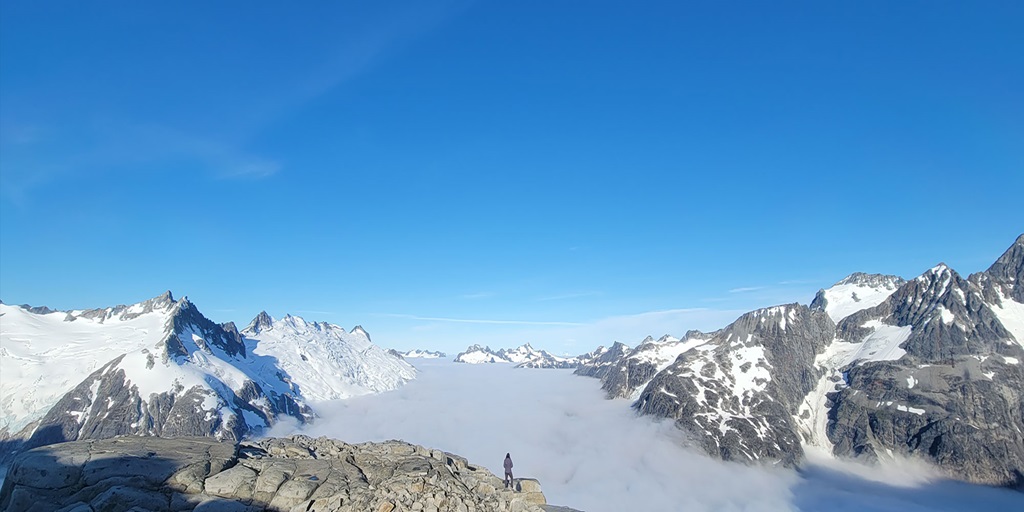 A person stands on a rocky cliff and stares out snow-capped mountains and clouds.