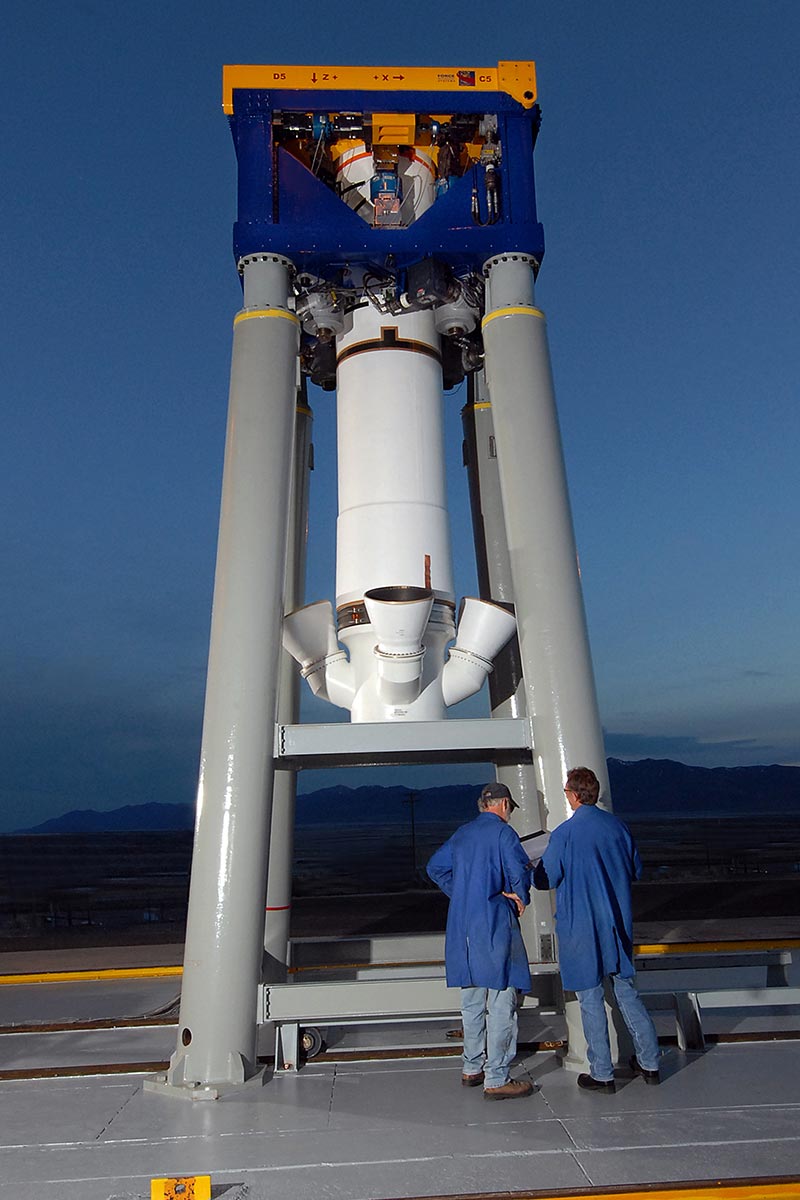 two men in blue coats stand in front of rocket test motor