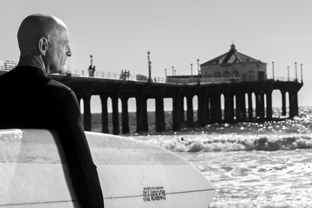 Older Caucasian man in wetsuit holds surfbboard at the beach