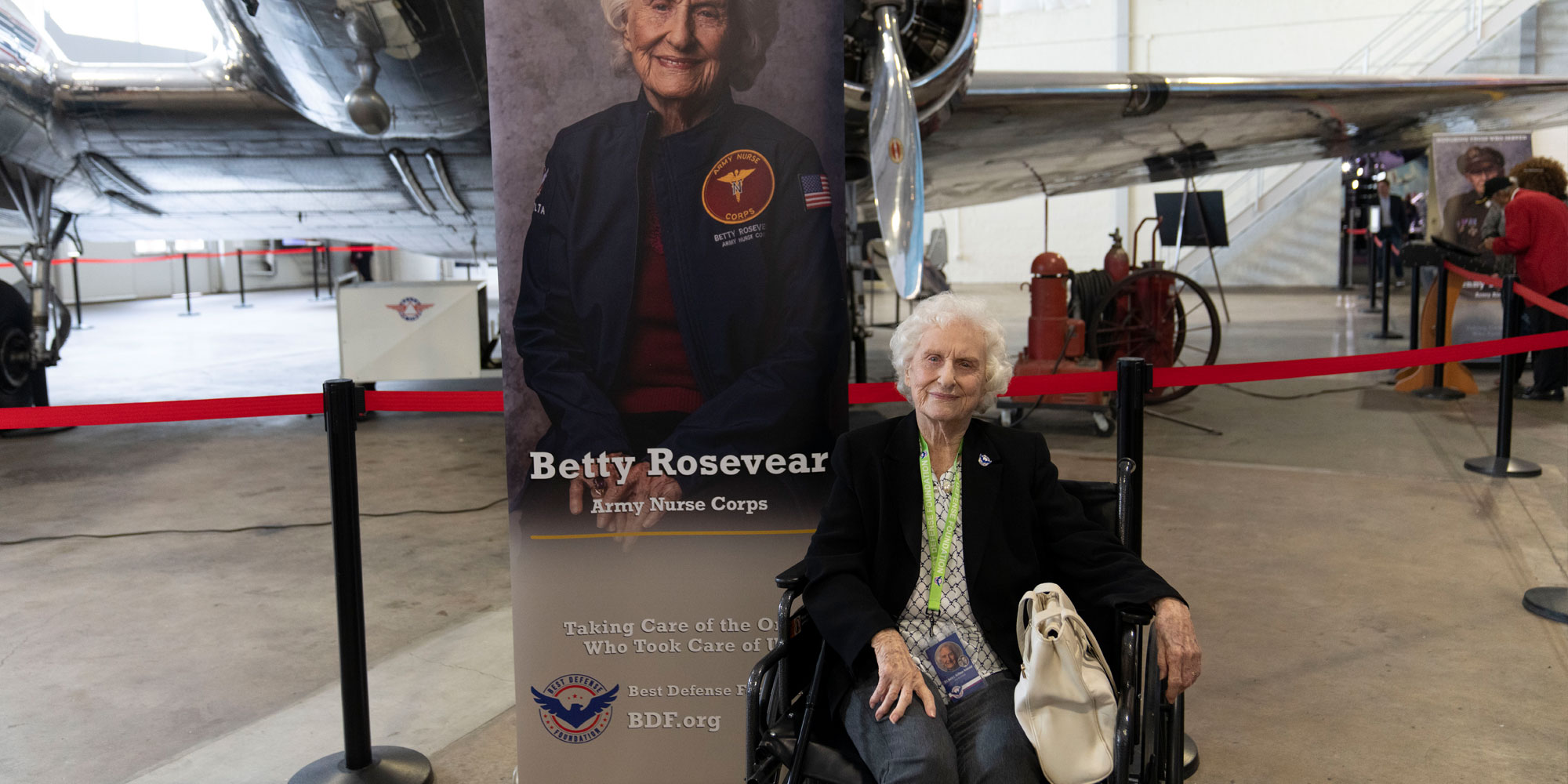 An older female veteran in a wheelchair sits in front of a plane and a poster featuring her