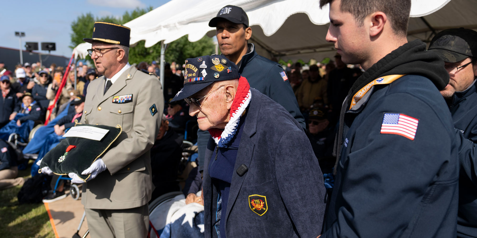 A group of male veterans and active duty military, including French military, at an outdoor ceremony. An older male veteran, wearing a red, white and blue necklace and ball cap, stands to receive his honor