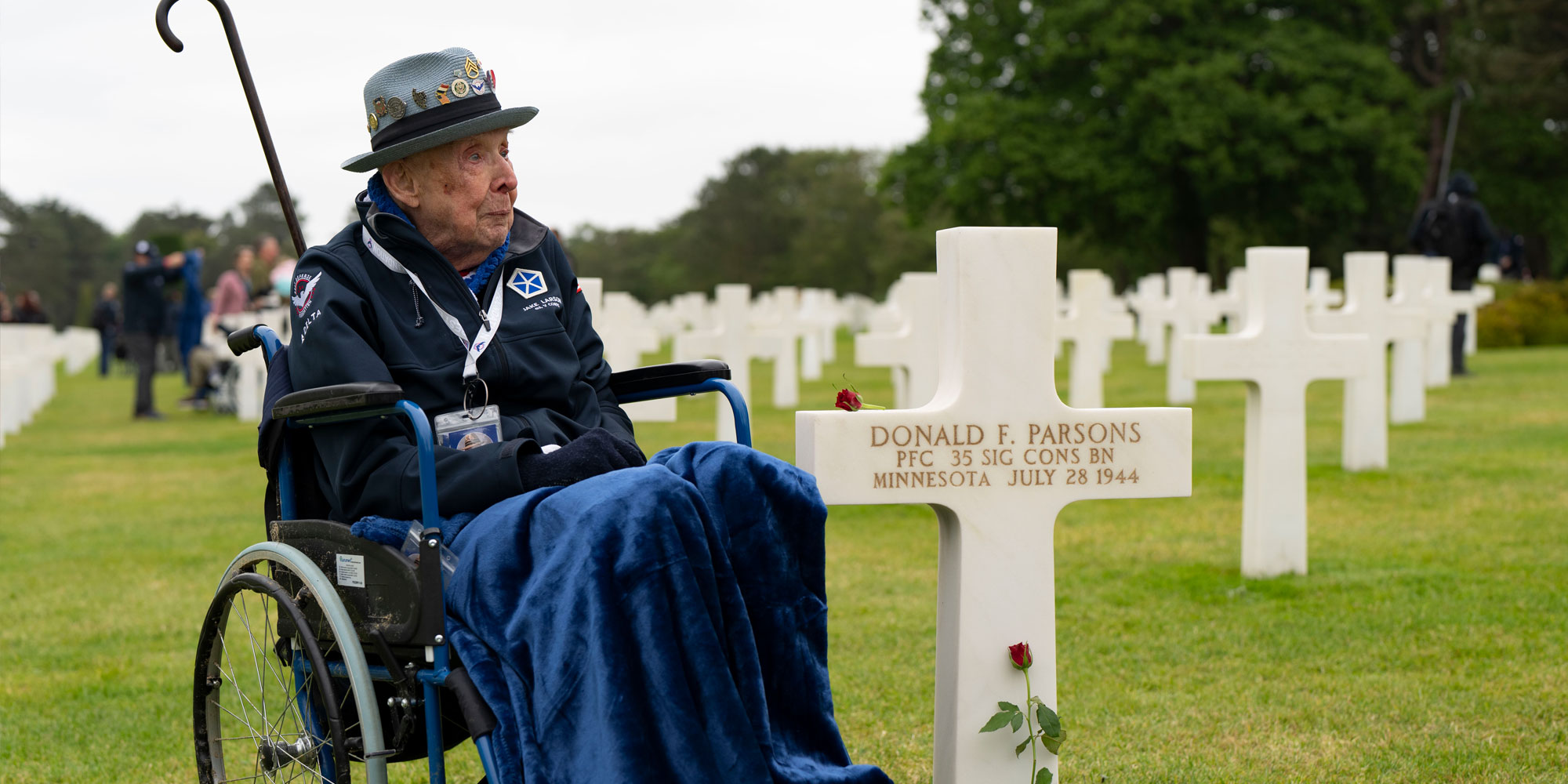 An older male veteran sits in a wheelchair, with a blue blanket on his lap, in a gravesite. He is next to a white marble cross tombstone with a single red rose leaning against it