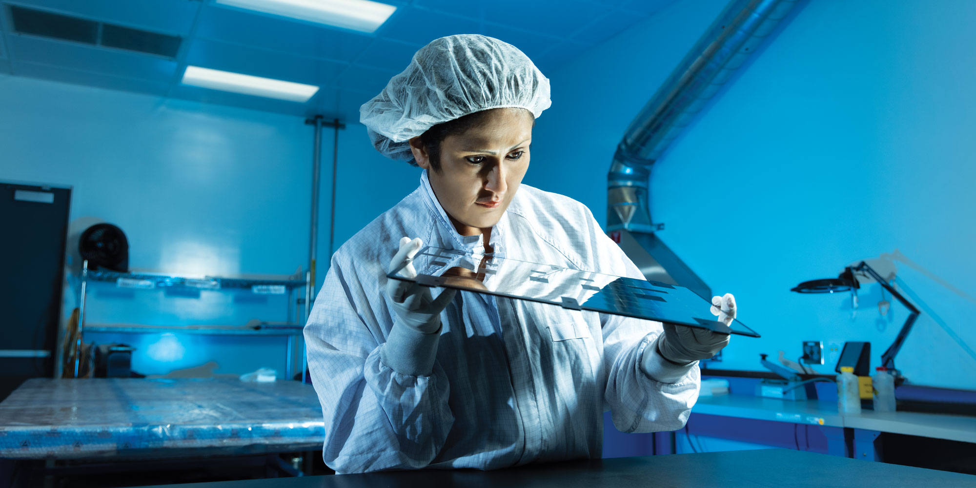 Rosibel Diaz wears a white coat, gloves and hairnet and stands at a table in a blue-lit lab, examining a mirrored panel in her hands.
