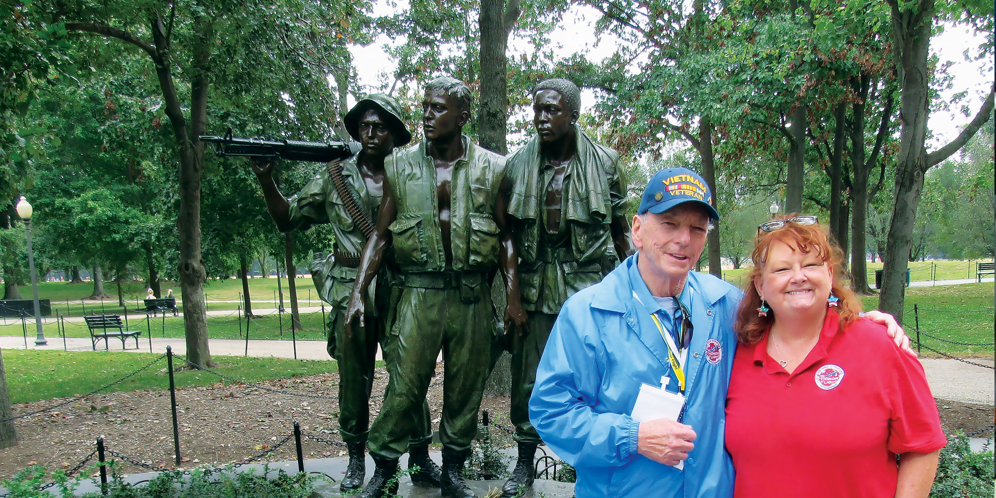An older man wearing a blue Vietnam veteran hat and jacket poses with a red-headed woman in a red Honor Flight Network shirt in front of a green and black memorial statue of three Vietnam soldiers.