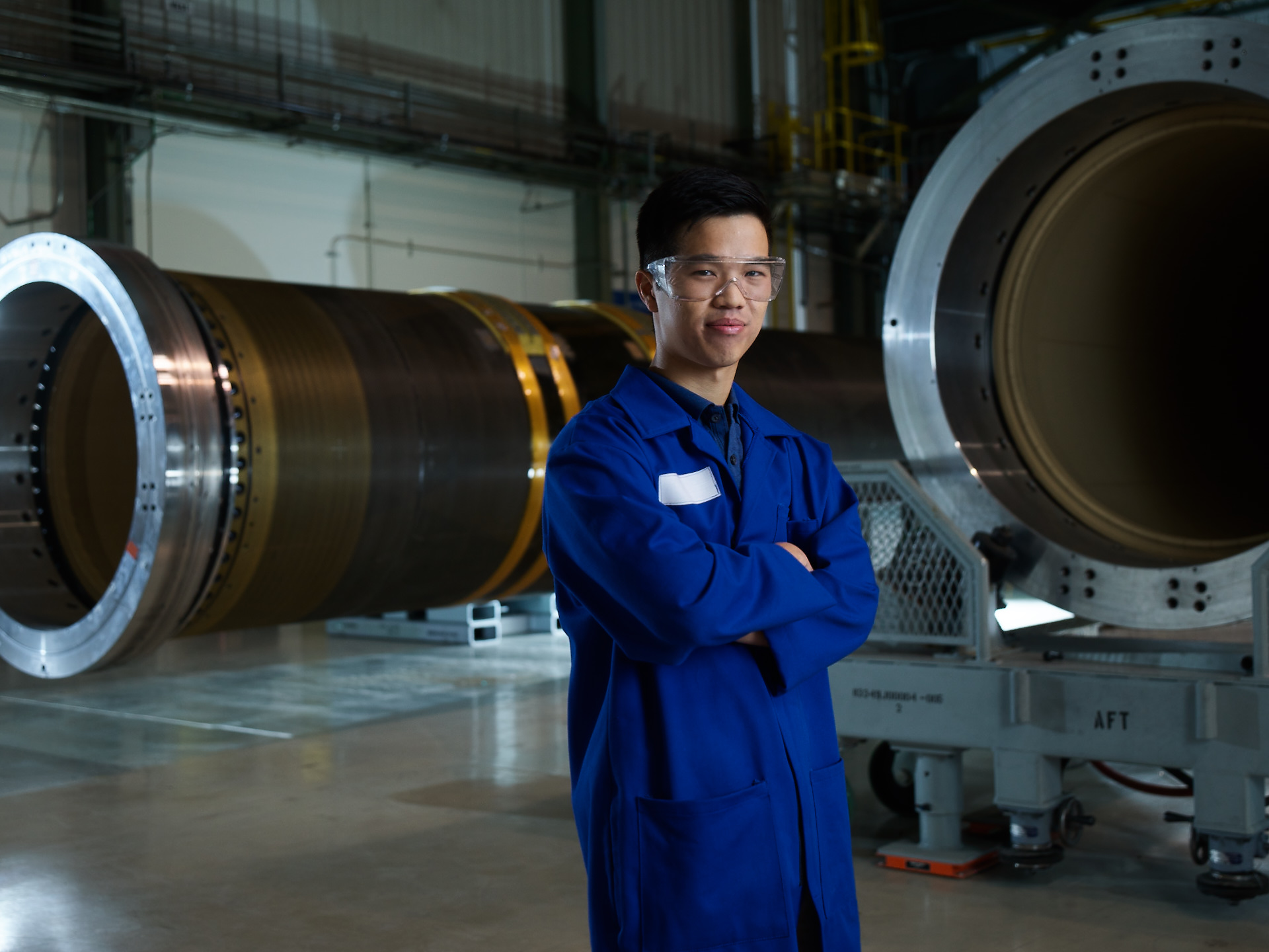 Asian male in blue lab coat and safety goggles stands in manufacturing plant in front of rocket motor frames.