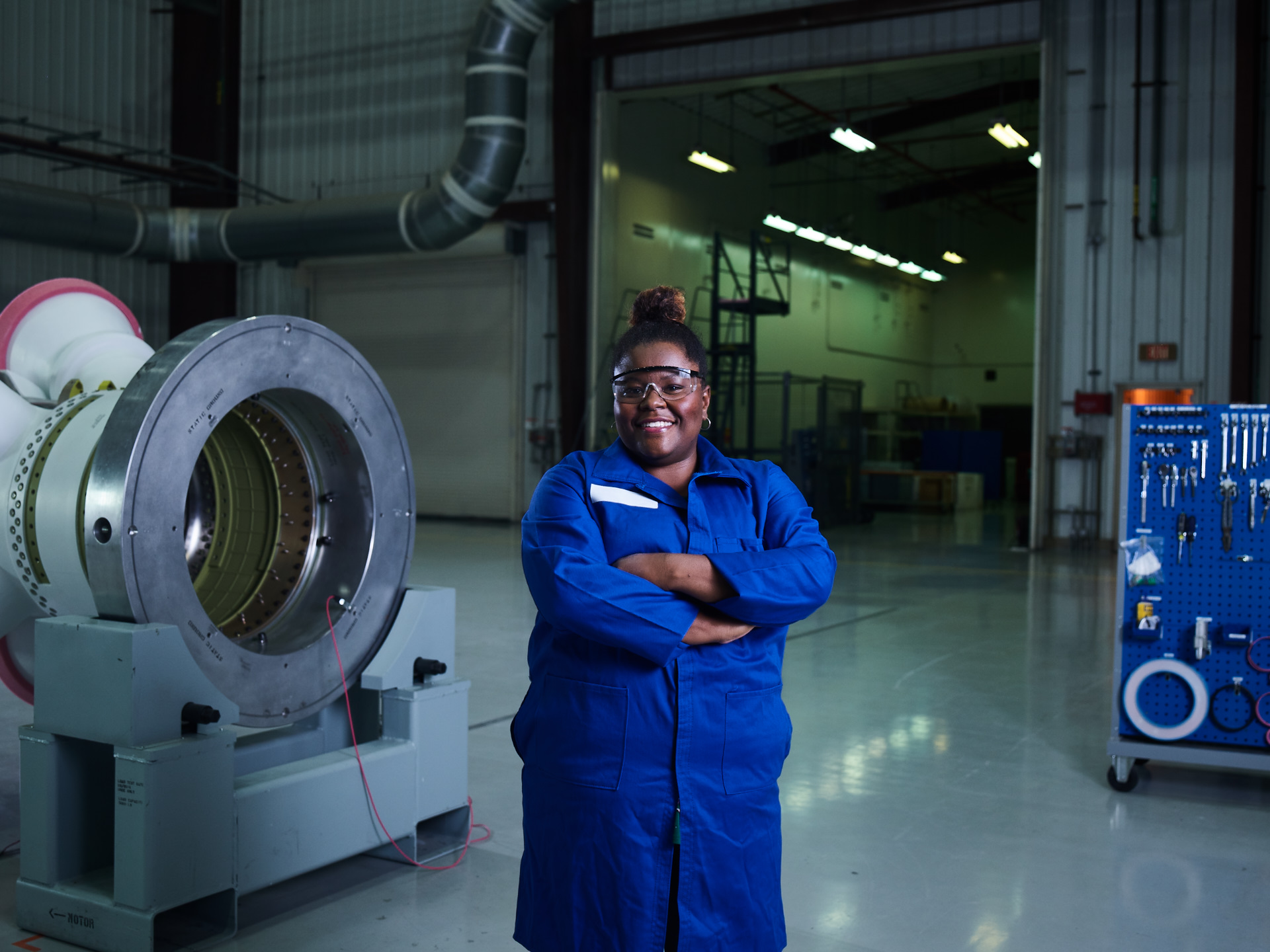 Black female in blue lab coat and safety goggles stands in manufacturing plant in front of rocket motor frame.