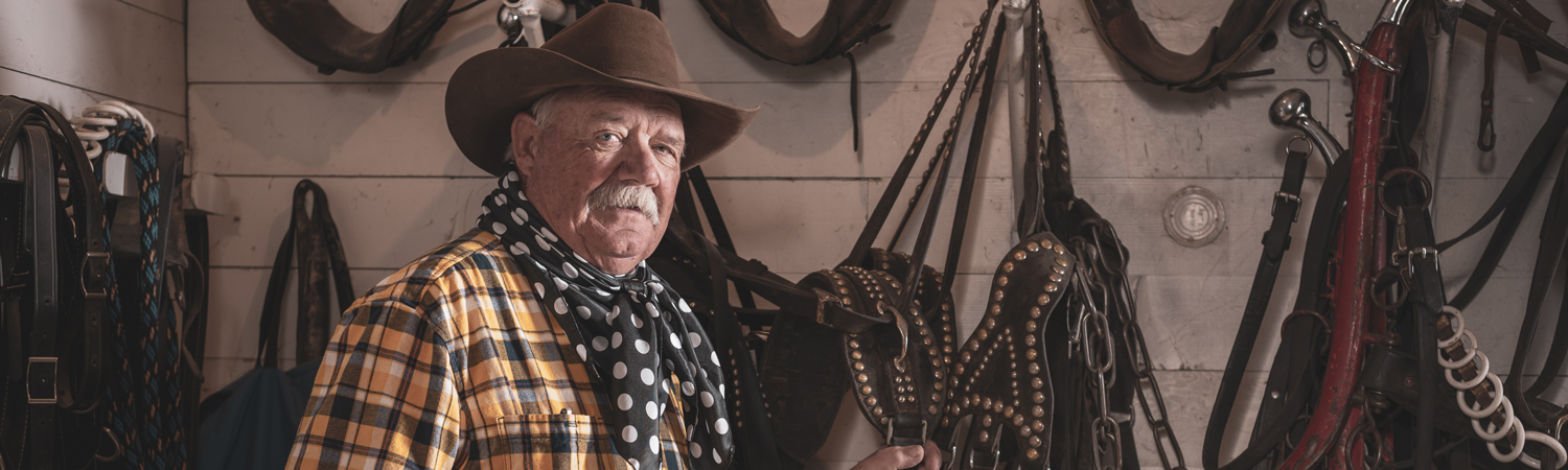 Older Caucasian man in cowboy attire poses in front of horse saddles hanging on wall