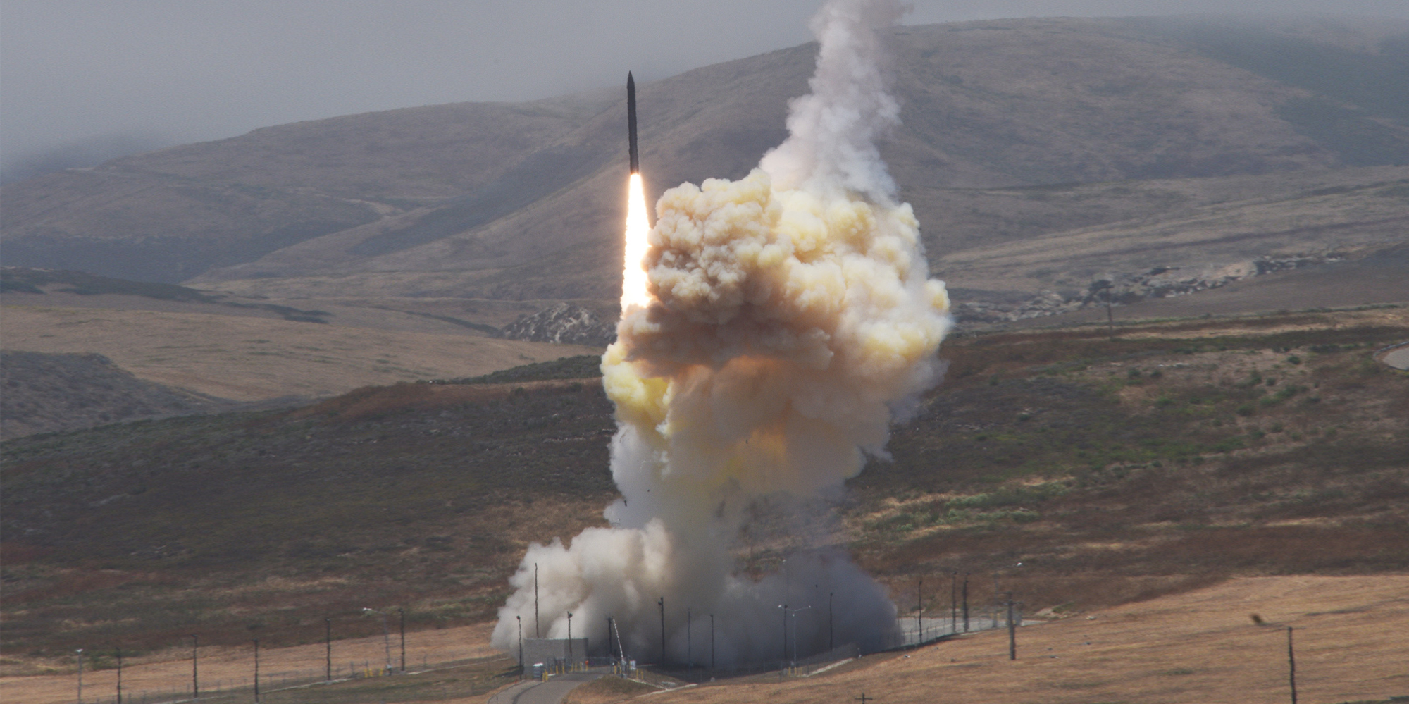 A missile interceptor launching from test field in to a gray sky in front of mountains