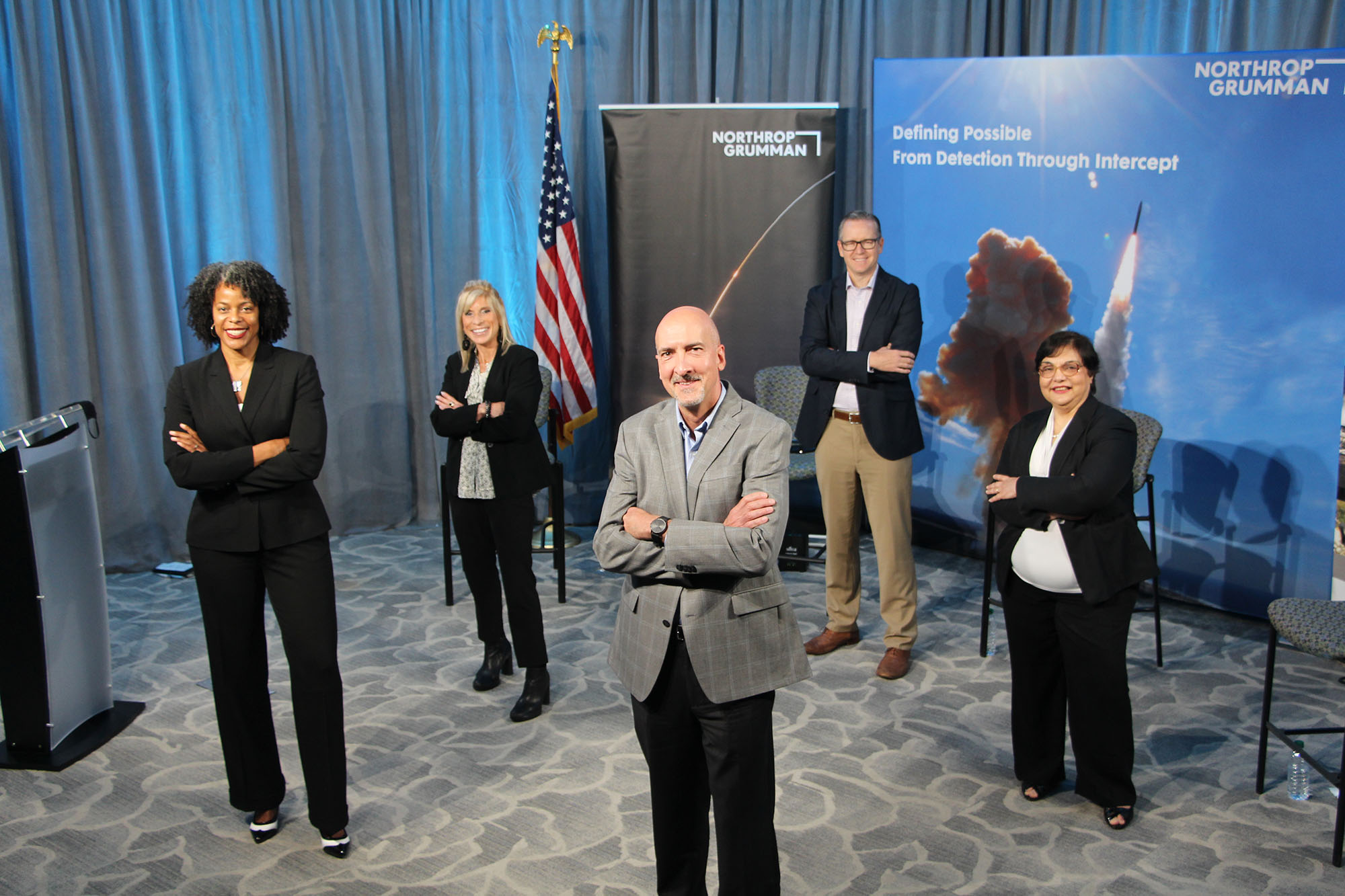 From left to right: A black female, white female, white male, white man and hispanic lady pose for a picture at a townhall in front of an American flag and company back drop