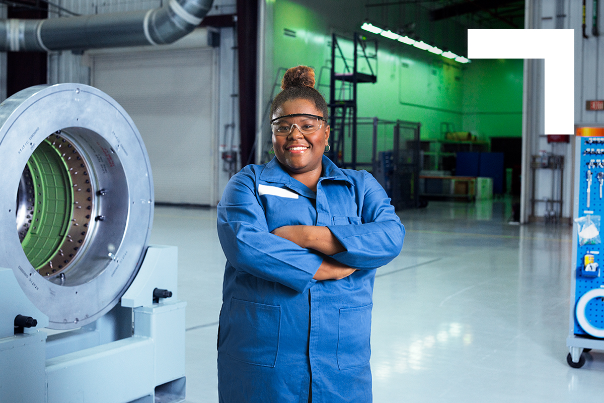 African American female wearing blue jumpsuit posing with arms crossed in front of production plant